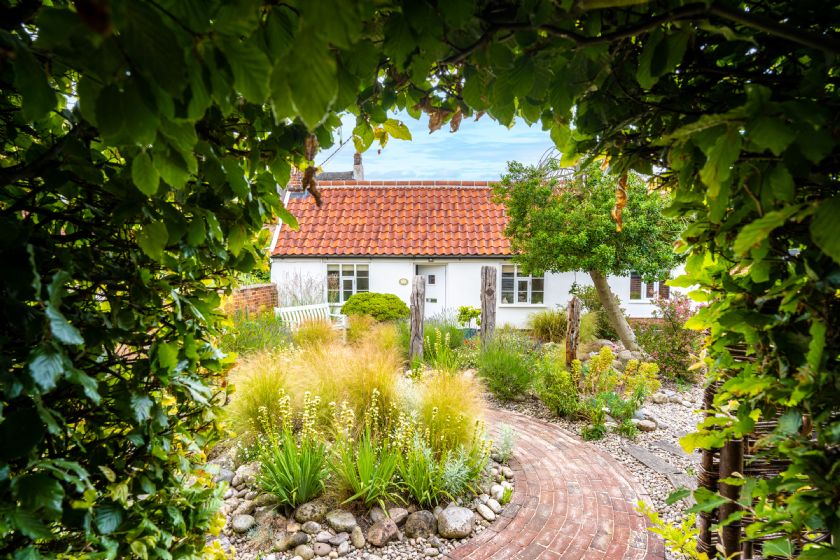 White cottage with terracotta roof seen through leafy archway with winding brick path